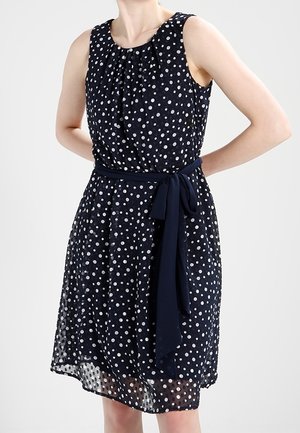 Granatowa sukienka bez rękawów w białe groszki, z talią podkreśloną pasem z tego samego materiału zawiązanym na kokardę.