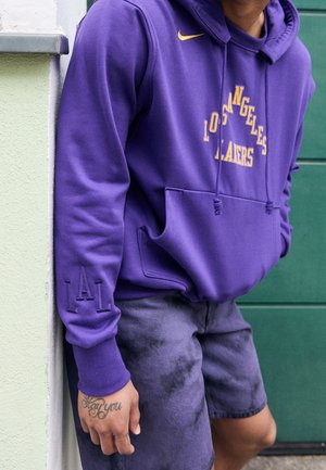 Osoba w fioletowej bluzie z kapturem Los Angeles Lakers i ciemnofioletowych szortach, opierająca się o jasnozieloną ścianę z tatuażem na ręce.