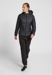 Zwarte pufferjack met capuchon, doorgestikt ontwerp, rits aan de voorkant en zijzakken, in combinatie met zwarte trainingsbroek en zwarte schoenen.