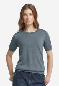 Kvinna med ljusbrunt hår iförd en kortärmad, grå stickad topp och mörkblå jeans, stående med händerna i fickorna mot vit bakgrund.