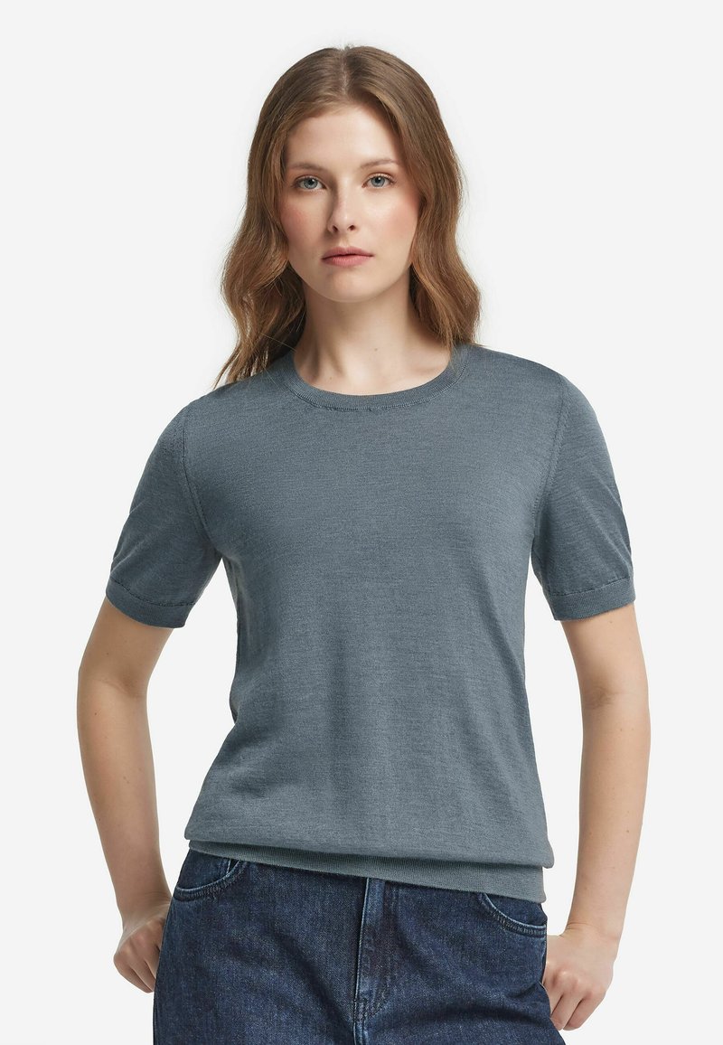 Kvinna med ljusbrunt hår iförd en kortärmad, grå stickad topp och mörkblå jeans, stående med händerna i fickorna mot vit bakgrund.