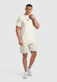 Camiseta atlética de color beige claro con cuello redondo y mangas cortas, combinada con shorts a juego. Presenta un logo en la camiseta y en los shorts.