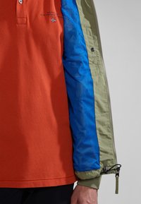 Polo rosso a maniche corte con colletto, abbinato a una giacca mista blu e verde con chiusura a zip e accenti in rete.