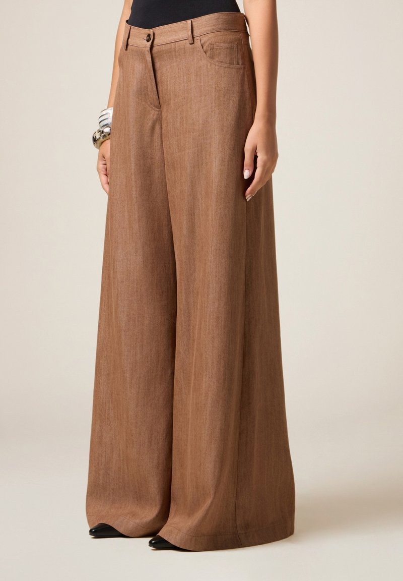 Pantalon large en tissu texturé de couleur marron, avec une fermeture à bouton, des poches avant et une silhouette fluide. Des chaussures noires pointues sont visibles.