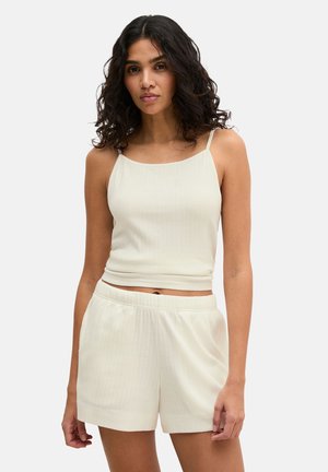 Donna con capelli ricci e scuri che indossa un top corto crema a coste senza maniche e pantaloncini crema abbinati, in piedi davanti a uno sfondo neutro.