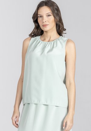 Kobieta o falujących brązowych włosach, ubrana w bezrękawową bluzkę w jasnym miętowym kolorze i dopasowaną spódnicę, stojąca na tle jednolitego jasnoszarego tła.