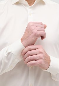 Weißes Langarmhemd mit glatter Textur, ausgestattet mit Knopfmanschetten. Hände passen die Manschette an und betonen die maßgeschneiderte Passform.