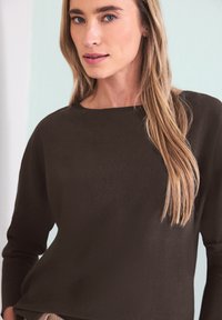 Femme aux cheveux blonds longs portant un haut à manches longues marron foncé, posant devant un mur de couleur claire.