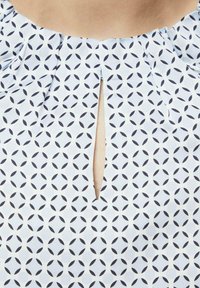 Hellblaue Bluse mit geometrischem Muster aus dunkelblauen Kreisen. Verfügt über eine Schlüsselaußenschnittöffnung und gefältelte Kragen-Details. Hergestellt aus glattem Stoff.
