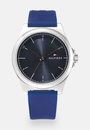 Runde silberne Armbanduhr mit schwarzem Zifferblatt, weißen Stundenmarkierungen, drei Zeigern, blauem Silikonarmband und Hilfiger-Logo auf der rechten Seite.