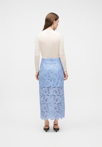 Γυναίκα με πλάτη, φορώντας μπεζ μπλούζα με μακριά μανίκια, φούστα MIDI από ανοιχτό μπλε δαντέλα με floral σχέδιο και μαύρα σανδάλια με χαμηλό τακούνι σε απλό φόντο.