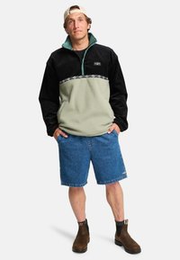 Black and green fleece pullover with a zip neck, featuring a geometric pattern on the chest, paired with blue denim shorts and brown boots.