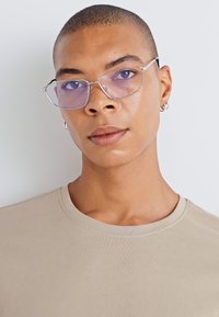 Des lunettes rectangulaires transparentes avec une fine monture en métal, des verres teintés en violet, et une chemise beige texturée dans un environnement neutre.