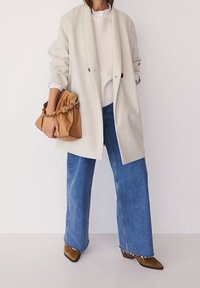 Blazer gris clair oversize sur un pull côtelé crème, associé à un jean large bleu et des bottines marron. Tenant un sac souple marron clair.