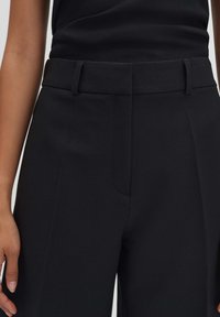 Schwarze, maßgeschneiderte Hosen mit glatter Textur, ausgestattet mit einem flachen Vorderteil, Gürtelschlaufen und einer verdeckten Reißverschlusstasche für ein elegantes Finish.