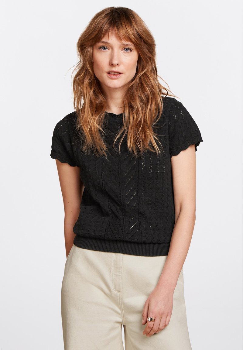 Schwarzes Kurzarmoberteil mit strukturiertem Muster und gewelltem Saum. Verfügt über einen runden Halsausschnitt und einen gerippten unteren Bund. Kombiniert mit beigen Hosen.