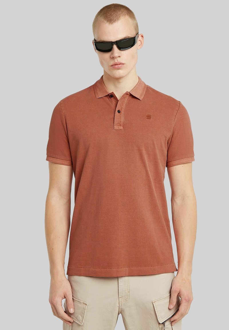 Polo shirt color ruggine con collo a polo, maniche corte, patta con due bottoni e un piccolo logo sul petto. Texture liscia.