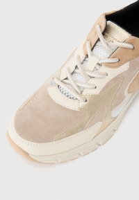 Beige semskinns sneakers med metalliske gull aksenter, teksturert mesh-detaling, hvite snøring, og en gummisåle med ribbet mønster.