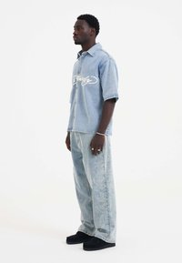 Blegnet denimskjorte med broderet hvid tekst, parret med lyseblå vidde jeans med indviklede blomstermønstre. Sorte sko fuldender looket.