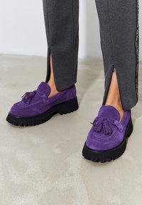 Lohkerit purppuranvärisestä mokasta, joissa on paksu musta kumipohja, koristeltuna hihnalla ja korostusompeleilla. Yhdistetty harmaisiin housuihin, joissa on sivuleikkaukset.