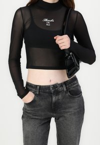 Vrouw in een doorschijnende zwarte lange mouwen crop top met de tekst "Thanks OK Bye", een zwarte tanktop eronder, vervaagde zwarte jeans en een zwarte handtas in haar hand.