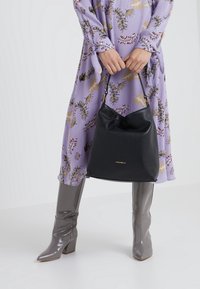 Musta nahkainen ämpärilaukku, jossa on tekstuuripinta ja kultainen logo, on henkilön käsissä, joka on pukeutunut kukalliseen liilaan mekkoon ja harmaisiin saappaisiin.