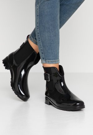 Piernas con jeans ajustados azules y botas de lluvia negras brillantes hasta el tobillo con detalle de hebilla sobre un fondo liso.