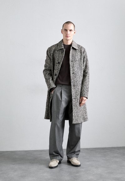 Pikk halli ja valge houndstooth muster särk, millel on lõige, kantud tumeda pruuniga kampsuni ja hallide plisseeritud pükstega, koos heledate jalanõudega.