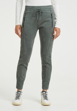 Pantalon de jogging gris charbon avec taille à cordon et poches latérales zippées, porté avec un haut blanc à manches longues et des baskets blanches.