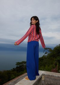 Femme aux longs cheveux foncés portant un pull rayé rouge et rose et un pantalon large bleu, debout sur un rebord en béton avec vue sur des arbres et de l'eau.