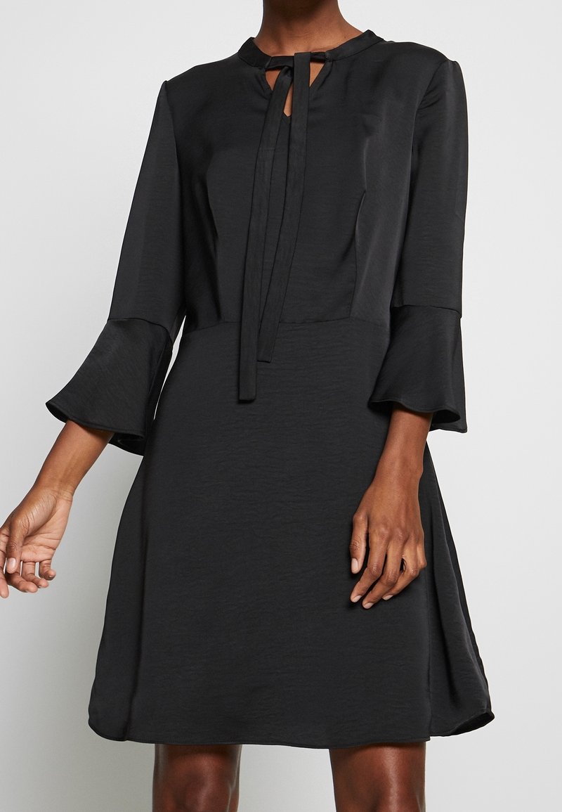 Robe noire jusqu'aux genoux avec des manches cloche 3/4 et un encolure en goutte d'eau nouée d'un petit nœud en tissu étroit au col.