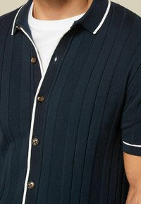 Navy lyhythihainen polo paita ryppypinnalla ja valkoisilla yksityiskohdilla. Sisältää napillisen kaulan ja ruskeat napit kiinnitystä varten.