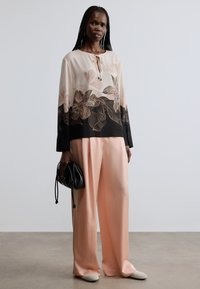 Blouse fluide à imprimé floral en rose pâle et noir, associée à un pantalon large couleur pêche clair. Complété par un sac à main noir et des chaussures douces.