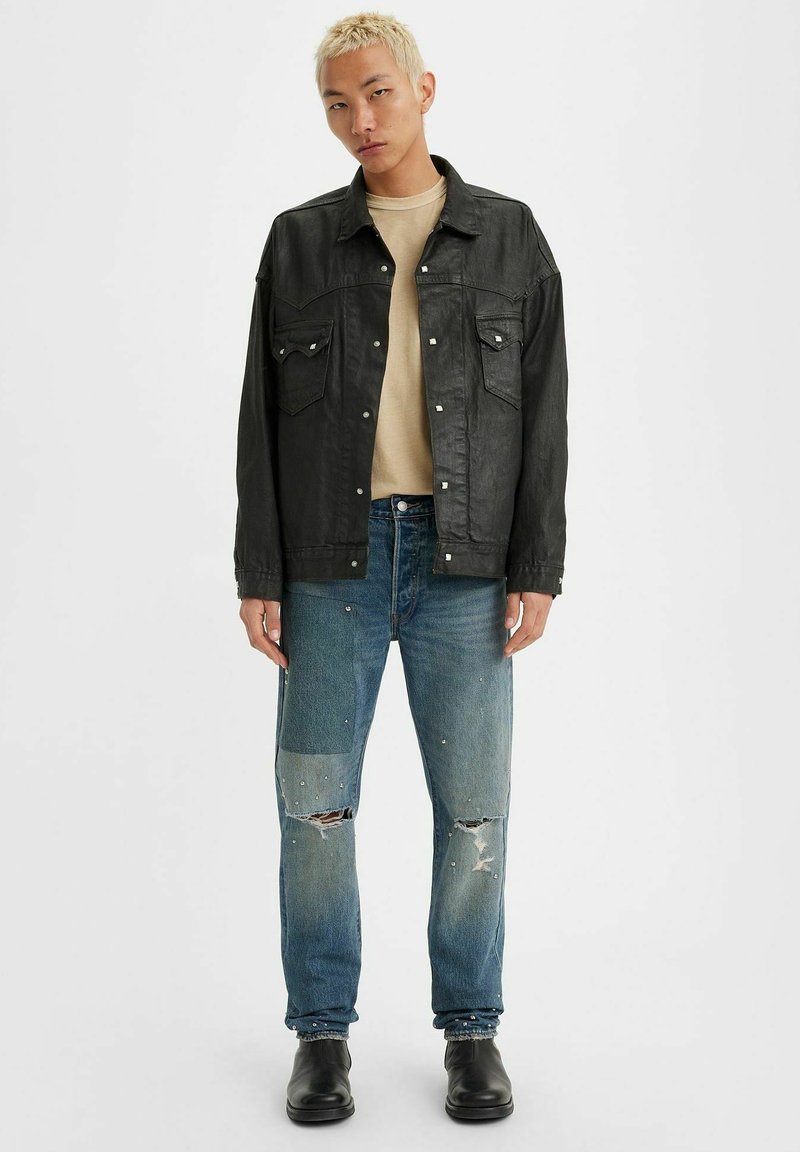 Chaqueta de piel sintética negra con dos bolsillos en el pecho sobre una camisa beige. Jeans azules desgastados con parches y botas negras hasta el tobillo.