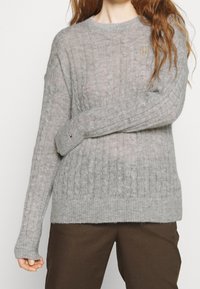 Pull gris à maille torsadée avec manches longues, col rond et ourlet côtelé. Présente un petit logo doré sur la poitrine.