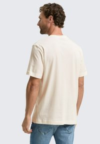Hombre con cabello rizado que lleva una camiseta corta de color blanco sucio y jeans azules, de espaldas frente a un fondo claro.