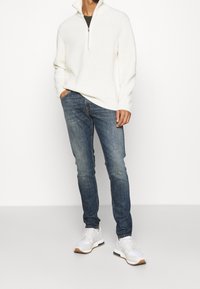 Pull blanc côtelé avec col à fermeture éclair quart, associé à un jean bleu ajusté et des baskets blanches avec des accents couleur caramel.
