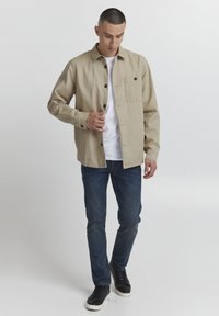 Beige langermet skjorte med knapplukking, brystlomme, brukt over en hvit T-skjorte, sammen med blå jeans og svarte sneakers.