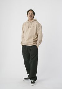 Beige Kapuzenpullover mit Fronttasche und Kordelzug, kombiniert mit schwarzen, lockeren Hosen und schwarzen Sneakers, vor einem weißen Hintergrund.