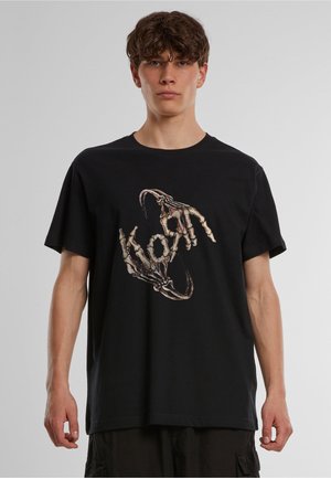 Ung mand iført en sort t-shirt med en grafik af to skelet hænder, der danner en cirkulær form på en ensfarvet baggrund.