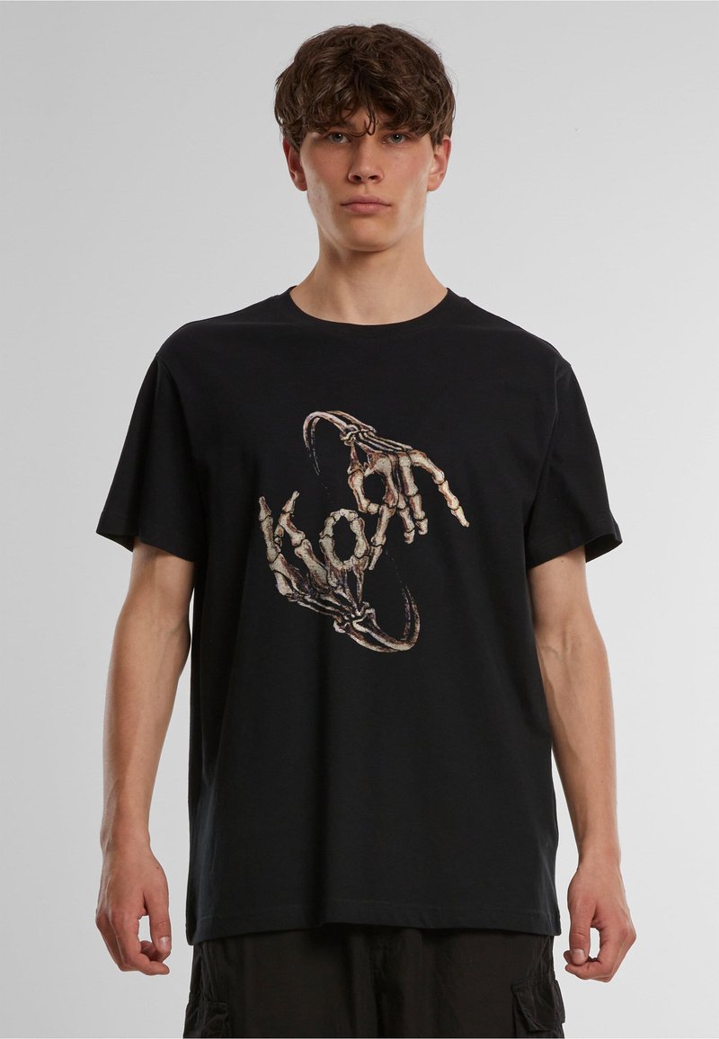 Jeune homme portant un t-shirt noir avec un graphisme de deux mains squelettiques formant une forme circulaire sur un fond uni.