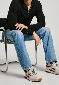 Polo a maniche lunghe nera con zip, jeans dritti blu chiaro e sneaker beige con strisce rosse e blu navy, seduto su una sedia cromata.