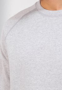 Primo piano di una persona che indossa un semplice maglione grigio chiaro a collo rotondo, con dettagli di cucitura visibili sulla spalla e sul collo.