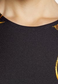 Černý top s texturou, který má zaoblený výstřih a výrazné žluté akcenty na ramenech, předvádějící hladkou strečovou tkaninu.