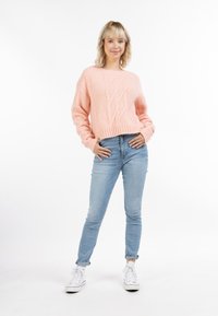 Rózsaszín, kábelmintás, rövid fazonú pulóver hosszú ujjakkal, világoskék skinny farmernadrág és fehér sportcipő fölött, fehér háttér előtt.