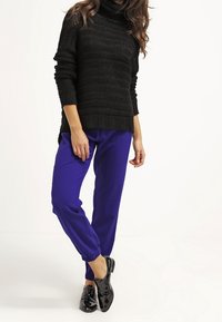 Maglione nero a maglia con un design testurizzato, abbinato a pantaloni tapered blu royal. Le scarpe sono mocassini neri lucidi.