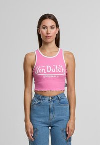 Jauna sieviete ar garām brūnām matiem, ģērbta rozā īsajā topā ar uzrakstu "Von Dutch Originals" un augsto jostasvietu zilos džinsos.