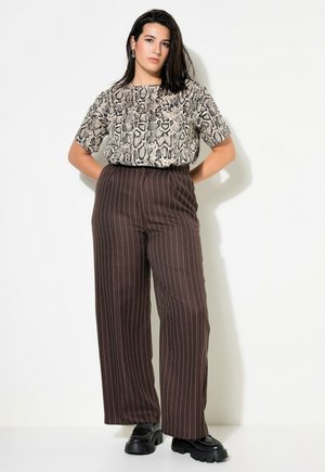 Una mujer posa sobre un fondo blanco con una blusa de estampado de serpiente, pantalones marrones de raya diplomática y pernera ancha, y zapatos negros de plataforma.