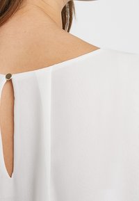 Weiße Bluse aus weichem Stoff, mit einem Schlüsselloch-Ausschnitt auf der Rückseite, der mit einem kleinen Metallknopf gesichert ist, und sauberer Naht entlang der Kanten.