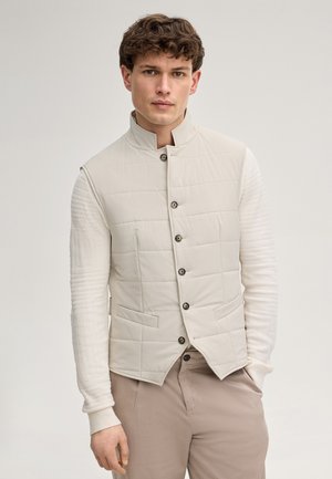 Mann trägt eine beige gesteppte Weste über einem weißen, strukturierten Langarmshirt und hellbraunen Hosen, steht vor einem neutralen Hintergrund.